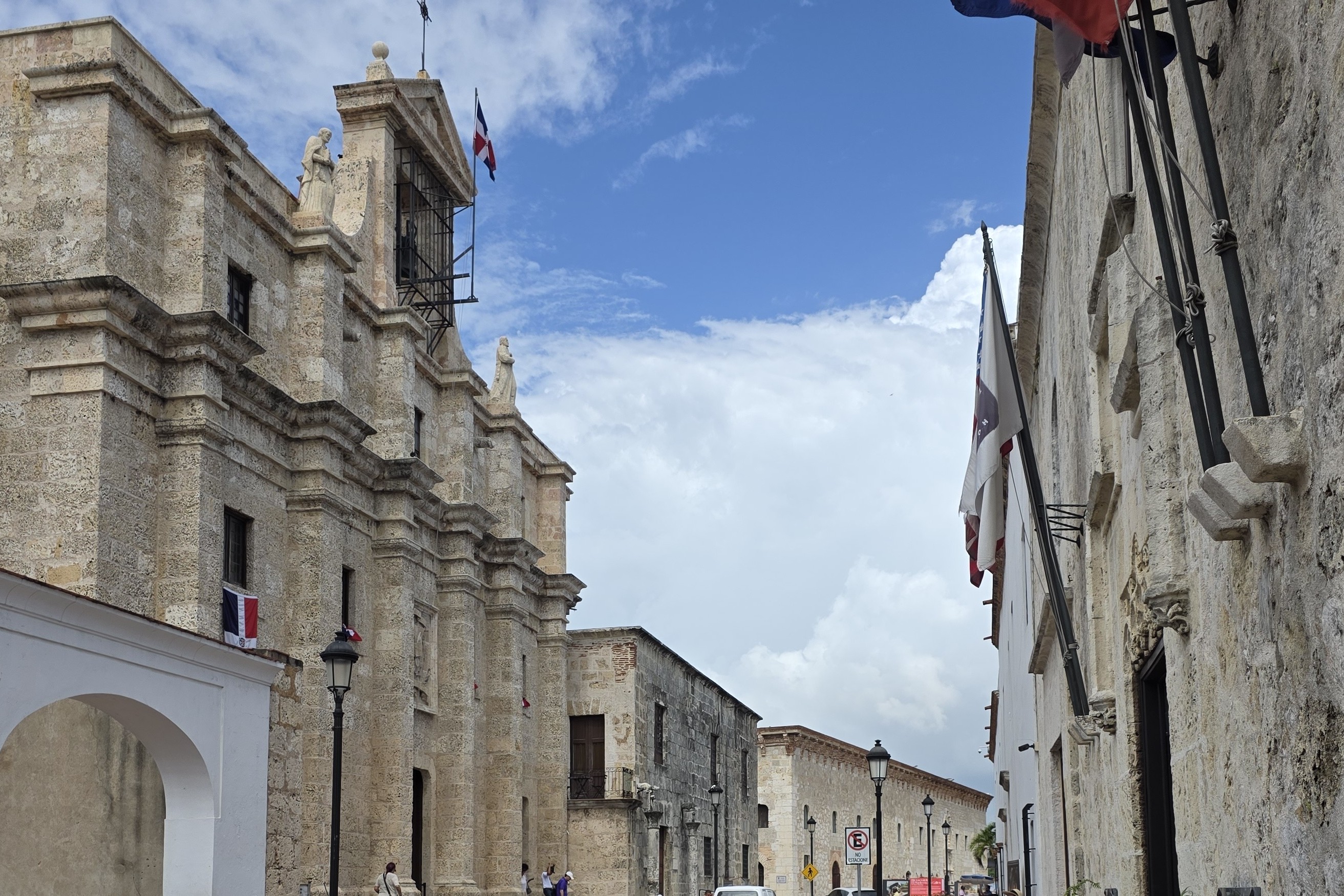 Calle Las Damas in the Colonial Zone of Santo Domingo, with historic stone buildings and Dominican flags under a cloudy blue sky