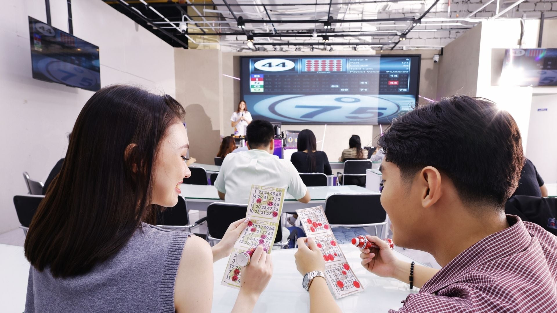 Players mark paper bingo cards in a BingoPlus hall, with a host and number display at the front.