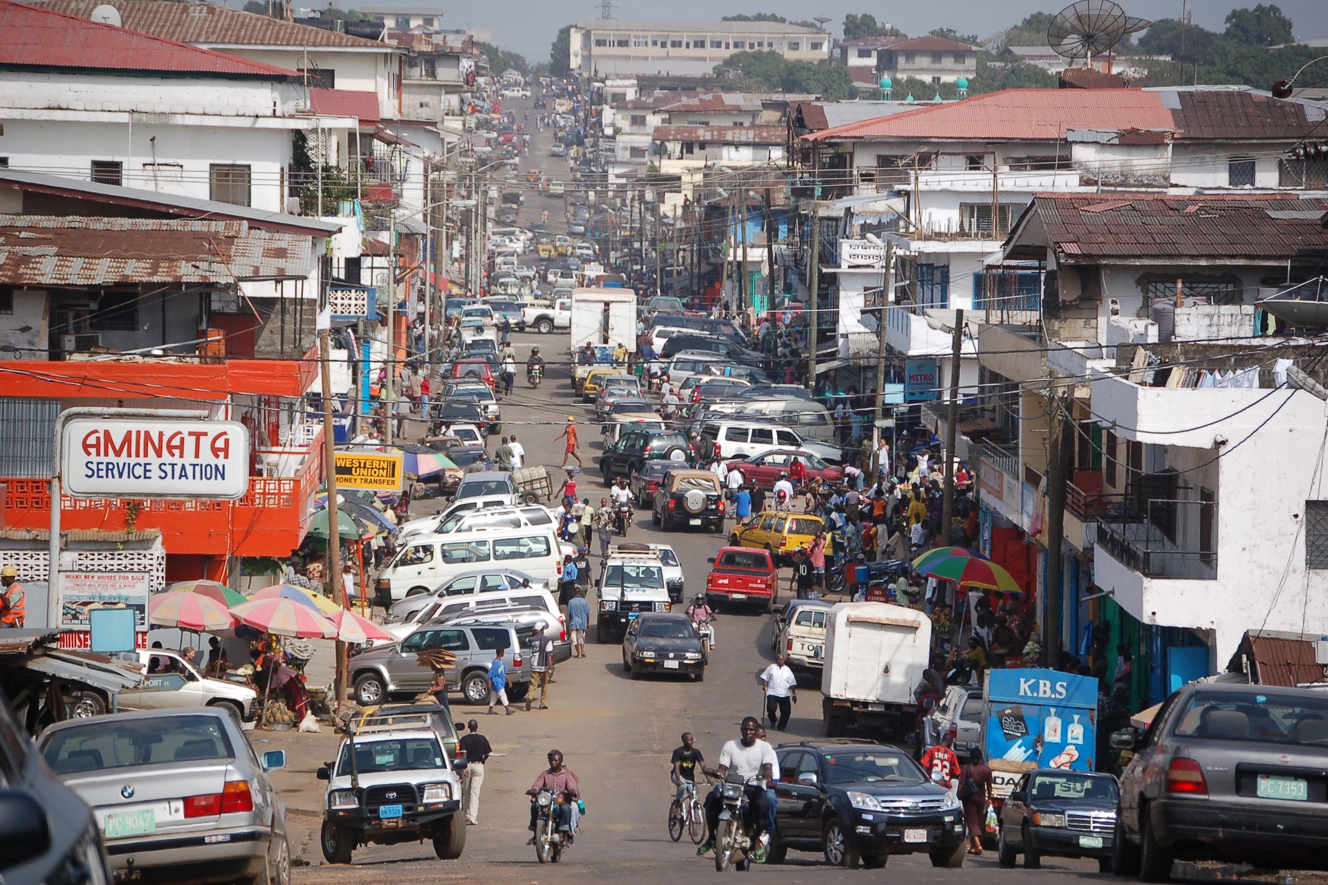 A busy downtown Monrovia street lined with shops, market umbrellas, cars and motorbikes, sloping down toward the city centre