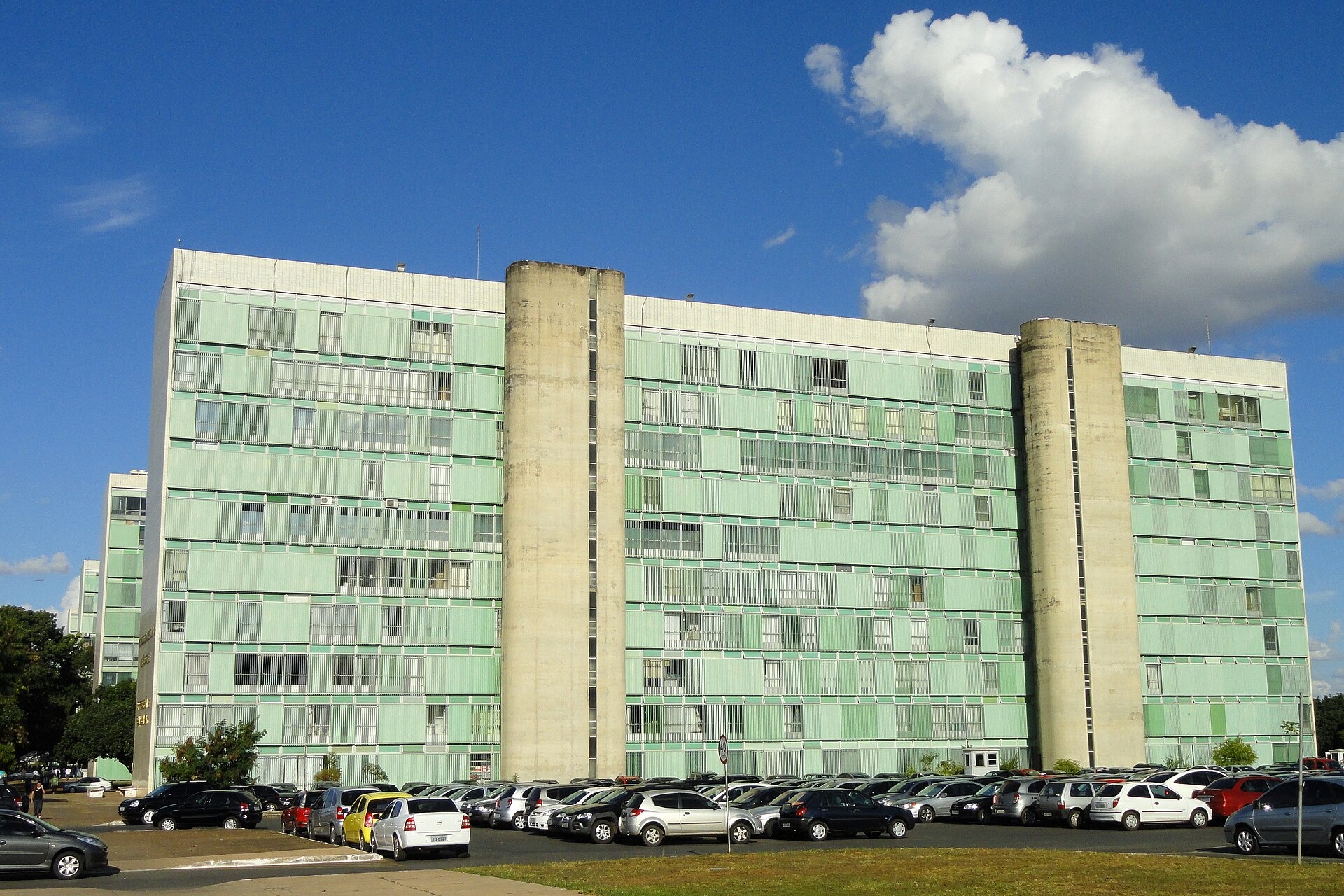 A green-glass government ministry building on the Esplanada dos Ministérios in Brasília under a blue sky with white clouds, cars parked in front