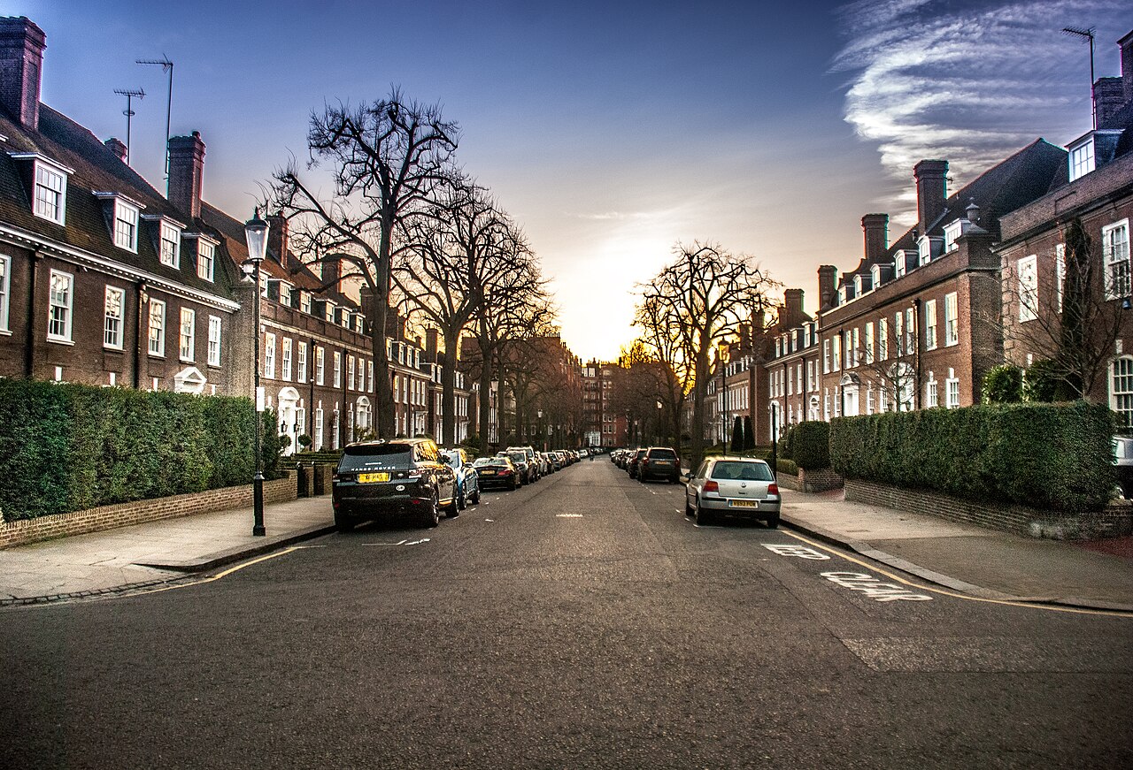 Tree-lined residential street in Kensington, London, with red-brick terraced houses and parked cars at sunset