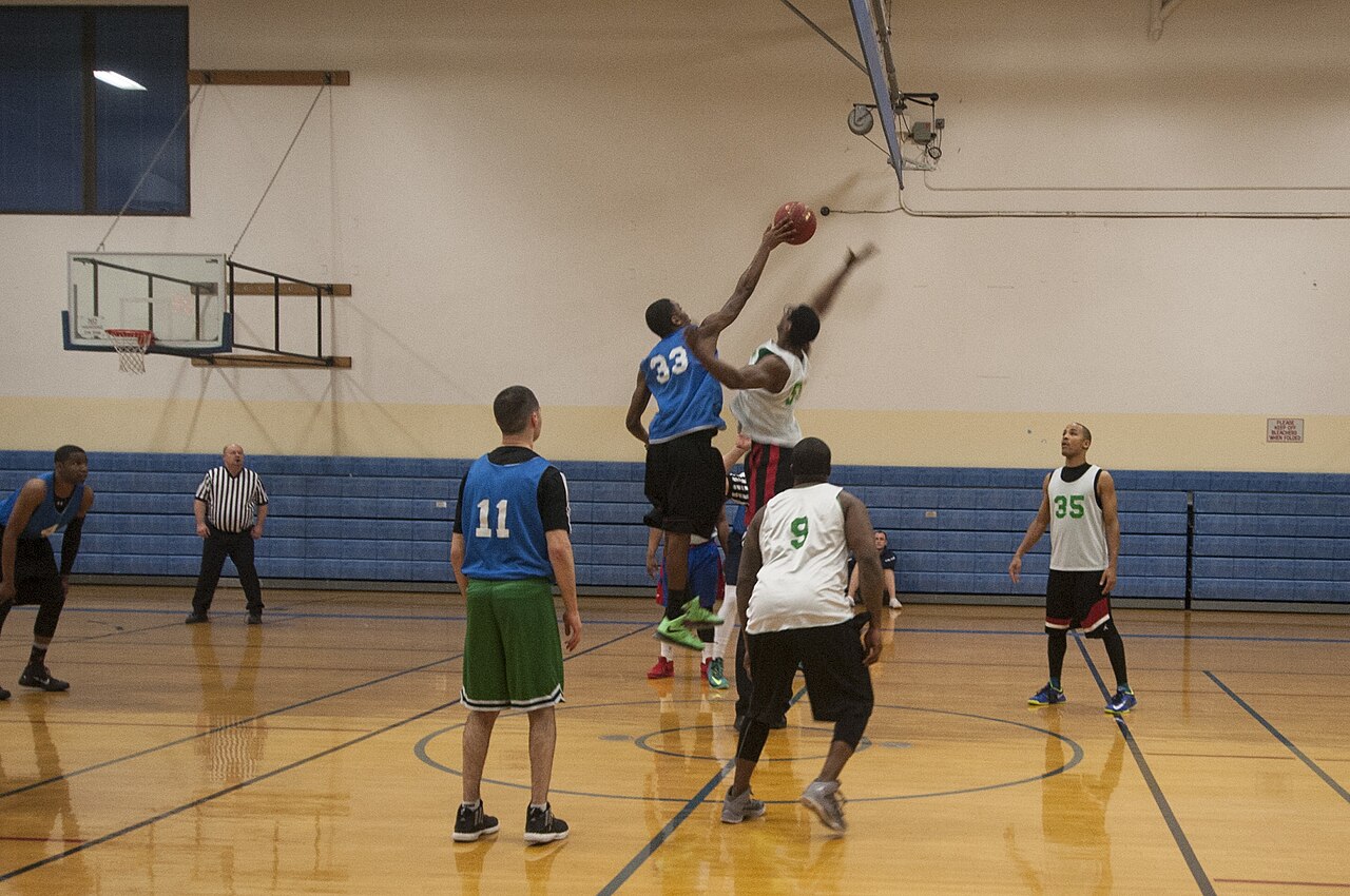 Two basketball players leap for the opening jump ball as a referee and other players watch in an indoor gym