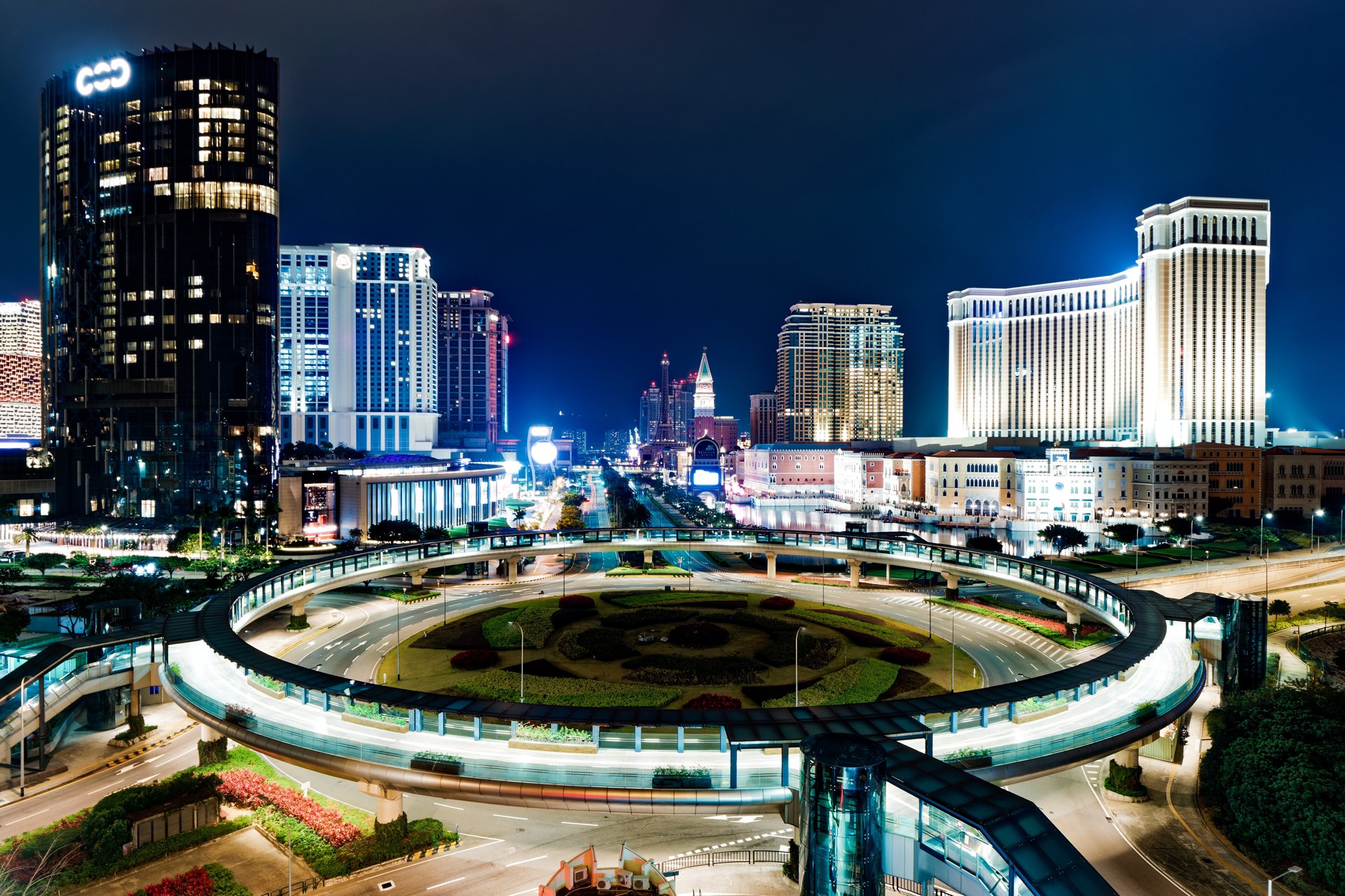 The Cotai Strip in Macau at night, with illuminated casino resort towers rising behind a circular elevated pedestrian walkway over a landscaped roundabout