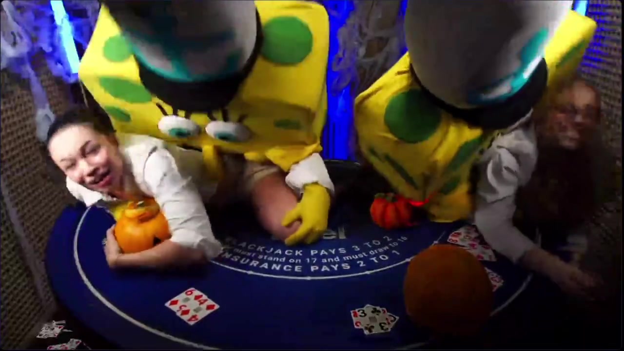 Two people in SpongeBob costumes lean over two dealers at a Duel blackjack table decorated with Halloween pumpkins