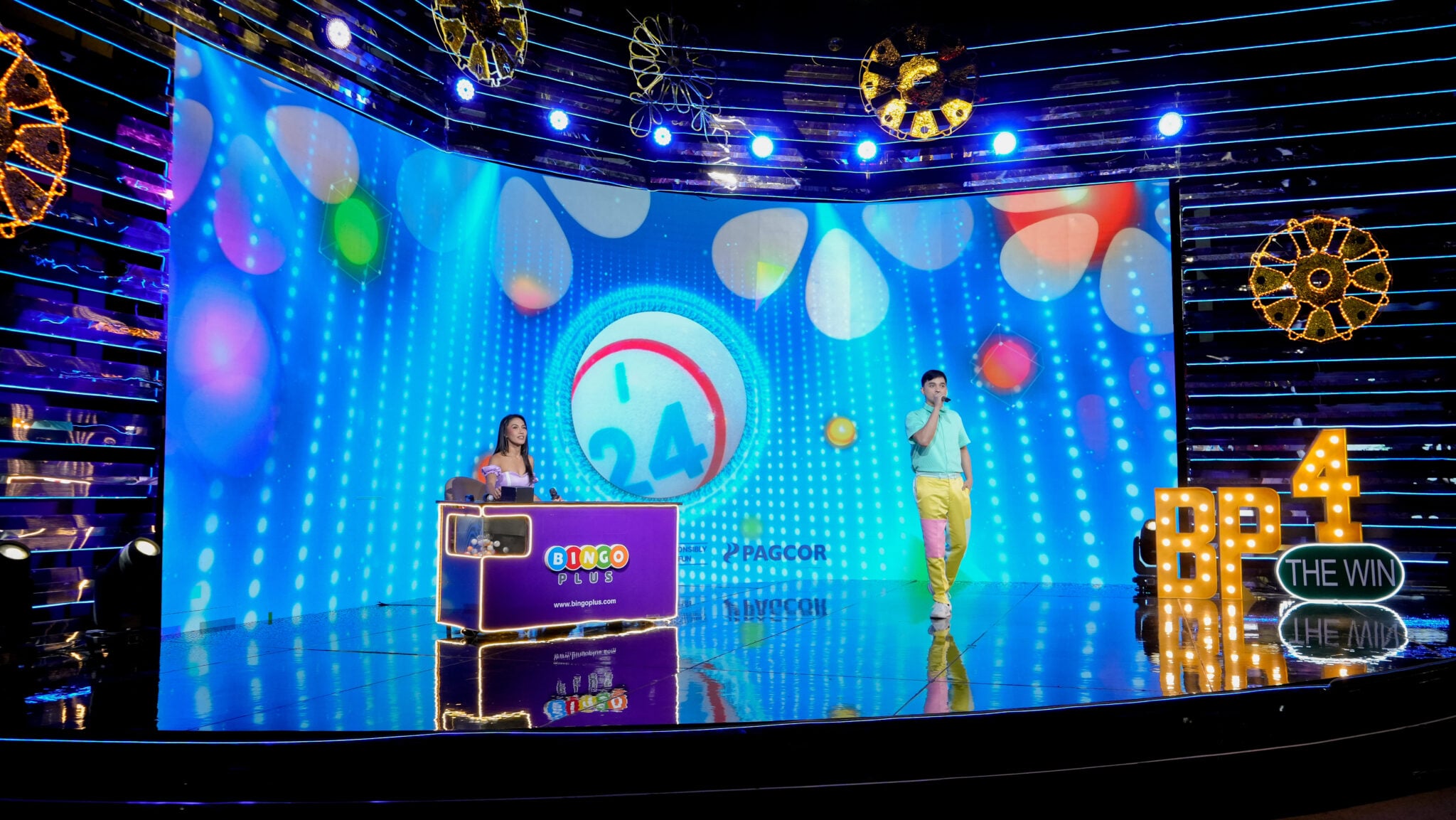 Two presenters in the Bingo Mega studio beside a BingoPlus-branded draw machine and a large numbered-ball display.