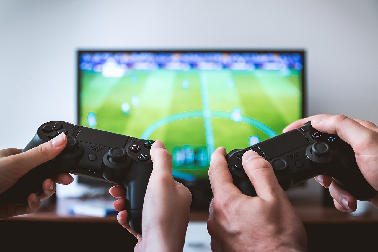 Two hands holding black game controllers in front of a television showing a football video game pitch