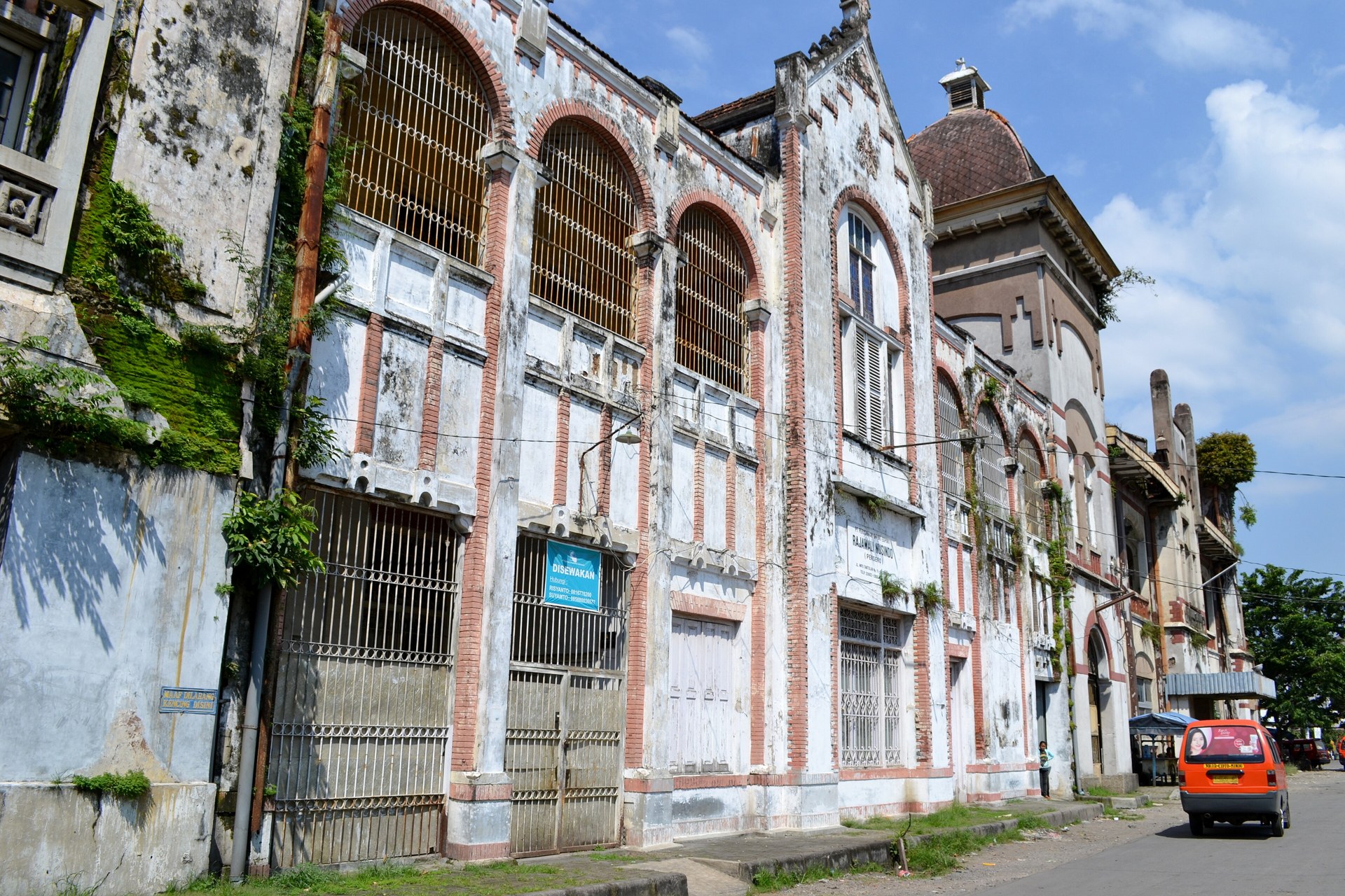 Weathered colonial-era buildings with arched, barred windows line a street in Kota Lama, the old town of Semarang, Central Java, as an orange minibus passes